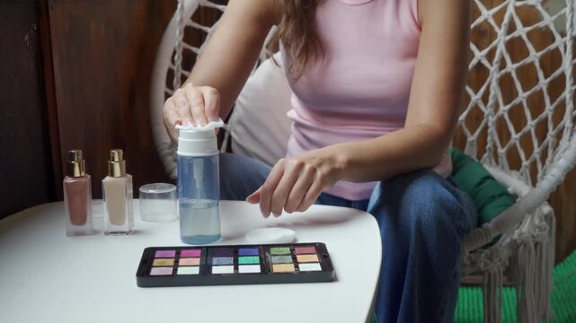 A focused woman gently presses a cotton pad onto a micellar water dispenser bottle. She happily prepares her skincare routine at a small white table inside a cozy balcony with a macrame chair.