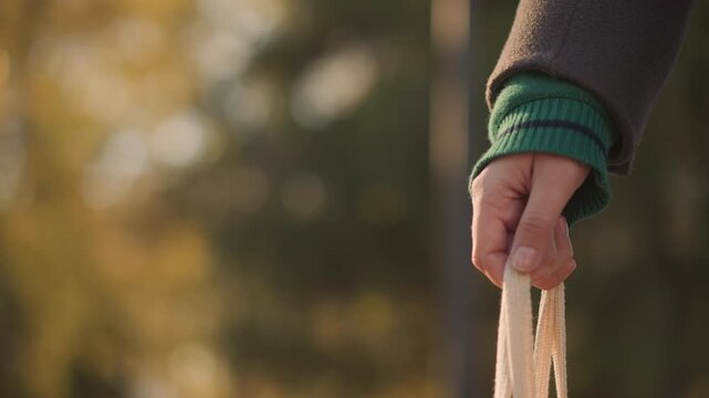 Close up of woman hand holding strap of off white bag with soft lighting and warm outdoor background. focus remains on grip detail throughout showing consistent positioning against blurred