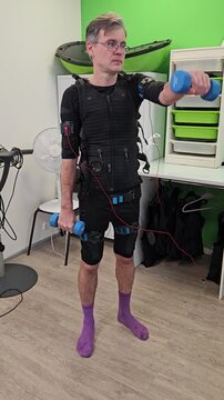 Man wears compression suit while lifting weights. He holds dumbbells, standing upright. He wears purple socks and glasses. Background includes green shelves and a fan electro muscle stimulation