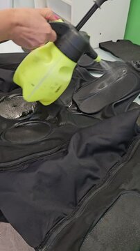 Person sprays black garment with green pump sprayer. Garment features padded sections and zippers. Scene shows pre-treating process. Action captured indoors on flat surface electro muscle stimulation