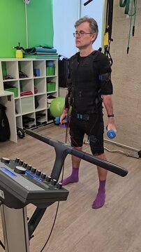 Man exercises with dumbbells in rehab facility. Wears support vest and purple socks. Treadmill nearby with control panel. Focused on movement with assistive tech electro muscle stimulation