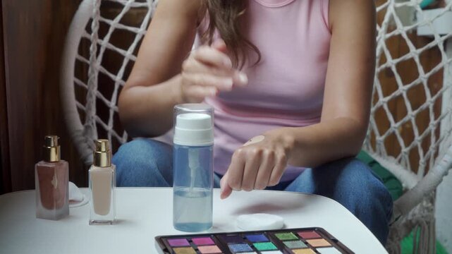 A focused woman gently tests a drop of liquid foundation on the back of her hand. She sits contentedly by a small white table with cosmetics inside a cozy balcony next to a macrame chair.