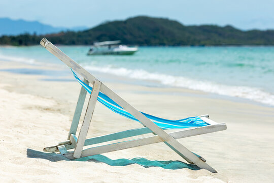 Wooden beach lounge chair with blue fabric rests on the sandy shore, with gentle ocean waves lapping nearby and a boat visible in the backdrop of hills. Summer tropical vacation and relax