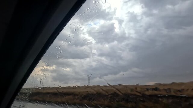 Storm clouds breaking beyond a rain-covered car window in side view during travel in Turkey, showing changing weather, wet road conditions, atmospheric contrast, regional mobility, and road trip