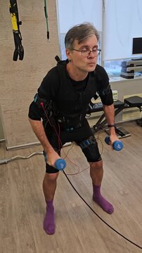 Man exercises using resistance gear indoors. He wears a black suit . He moves while gripping blue handles connected to a machine. He uses a body suit with wires for support electro muscle stimulation