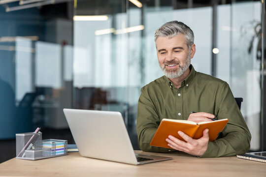 Confident senior businessman working in a modern office, smiling while watching his laptop and writing important notes in an open planner, focusing on strategy and planning for business success
