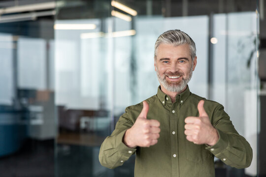 Happy mature businessman with grey hair and beard smiling, confidently giving a double thumbs up in a modern office environment, symbolizing agreement and achievement