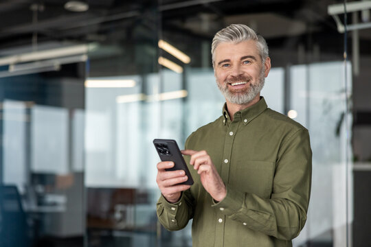 Confident mature businessman with gray hair and beard smiling at camera while using a smartphone, conveying modern communication, professional connectivity and success in a contemporary workplace