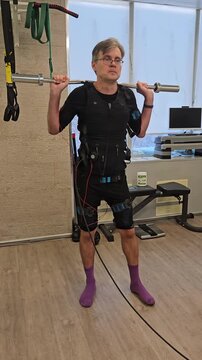 Man exercises with resistance equipment indoors. He wears a black suit . He holds a barbell while seated on a machine. His posture changes through controlled motion electro muscle stimulation