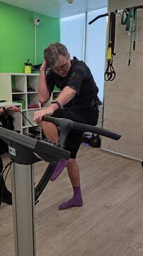 Man exercises on treadmill in rehabilitation facility. He wears purple socks and black athletic gear. He adjusts settings while holding his head. room fitness equipment and electro muscle stimulation