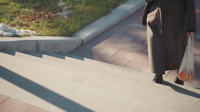 Close up of woman walking down steps outdoors carrying umbrella and net bag with fruit. Woman wears long coat boots against sunlit brick path backdrop. Soft shadows stretch across ground
