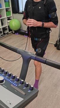 Man trains on exercise machine with vest and wires. He wears black gear . Equipment displays settings on control panel. Background shows green ball and storage shelves electro muscle stimulation