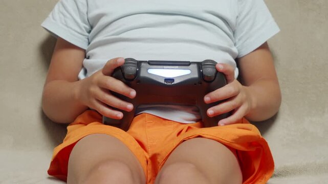 An excited child passionately holds a glowing black gamepad while playing video games. They sit cross-legged and focused on a comfortable, light-colored couch during a fun leisure routine at home.