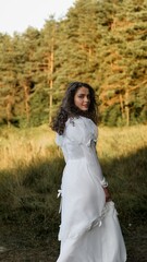 Fototapeta premium Young woman in a vintage white dress looking over her shoulder in a sunlit forest