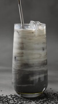 Metal straw stirs black sesame latte with ice, blending dark paste and milk into smooth marbled layers inside clear glass.