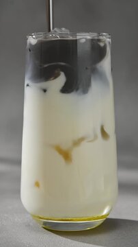 Black sesame paste is stirred into iced oat milk with metal straw, creating smoky marbled swirls in clear glass close up.