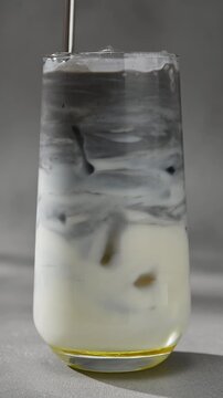 Metal straw stirs black sesame paste through iced oat milk, creating smooth gray marbled layers in clear glass close up.