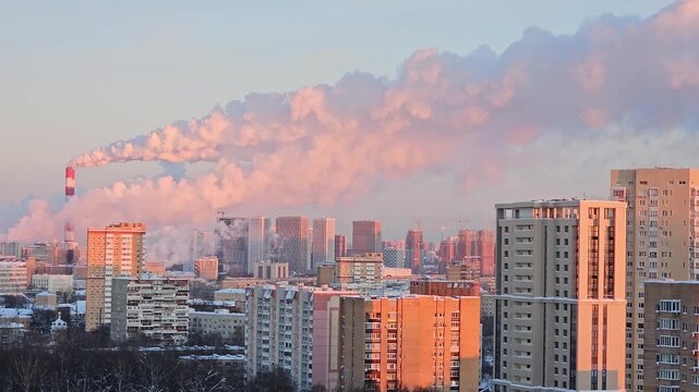 City skyline bathed in soft pink twilight. Smoke plume drifts from industrial chimney. Snow dusts buildings under hazy sky. Urban sprawl stretches into distant horizon
