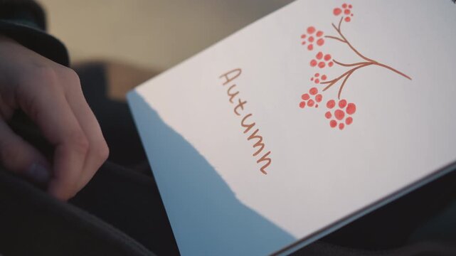 Close up of person holding and drawing on white paper with red berries illustration writing Autumn in cursive. hand holds sketchbook steady while using colored markers to add details