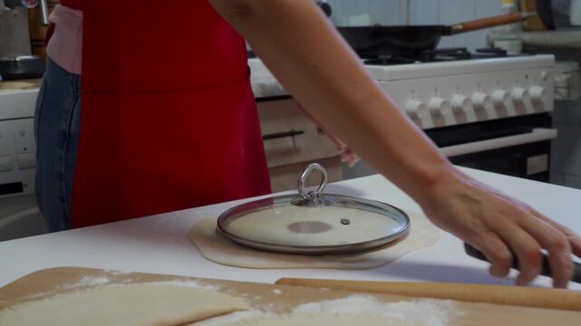 A woman peels excess dough away from a circular cut-out using a glass pot lid on a white table. She feels focused, enthusiastic, and content during her cozy home baking process.