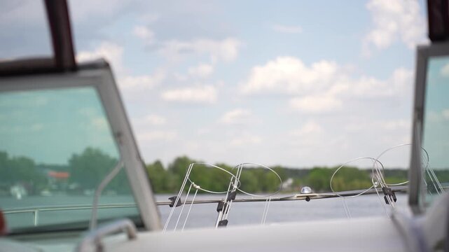 Boat bow rail with fishing rod guide loops framed by tinted windshields over calm lake under soft midday sky, with boat, rail, metal, fishing detail, showing rod, guide, holder, windshield context