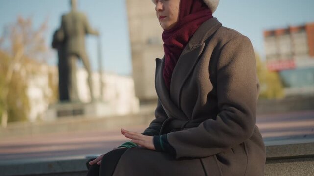 Close up of woman sitting on steps outdoors with hands resting wearing coat and scarf. Background includes statue and urban buildings under clear sky. Soft natural light creates warm tones
