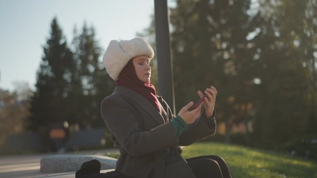 Woman in winter clothing takes selfie outdoors on bench with smartphone adjusting device and posing for photo against park backdrop with trees and sunlight