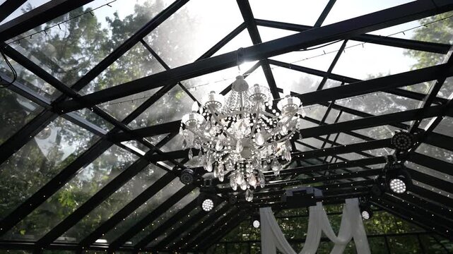 Crystal chandelier illuminated beneath glass roof with sunlit backlight and shimmering particles, with chandelier, crystal, backlight, sunlight detail, showing reflection, truss, glass, roof context