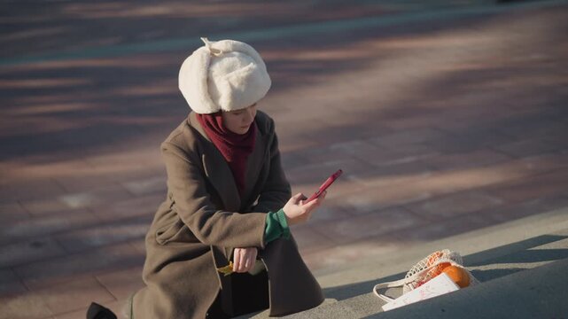 Close up of young woman sitting on steps outdoors holding and looking at her red smartphone. Woman wears white hat with ear flaps brown coat over green sweater maroon scarf next to her