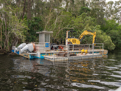 Yellow excavator works on floating platform near riverbank. Heavy machinery lifts pipes into water. Trees line riverbank