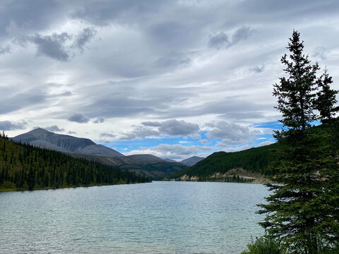 View of lake with mountains and evergreen trees under cloudy blue sky