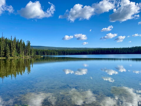 Calm, reflective lake near mountains and forest with white clouds in blue sky