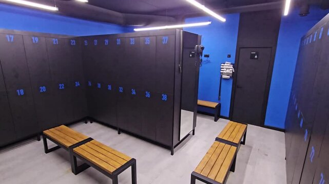 Modern gym locker room with blue walls and dark lockers. Wooden benches line the space under bright overhead lighting. Clean, organized room features numbered lockers and mirrors