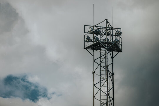Metal communication tower with floodlights against a cloudy sky, showcasing a tall structure with multiple light fixtures and a steel framework