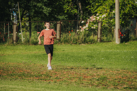 Young boy in orange t-shirt and black shorts runs on grassy field with trees and fence in the background during a sunny day