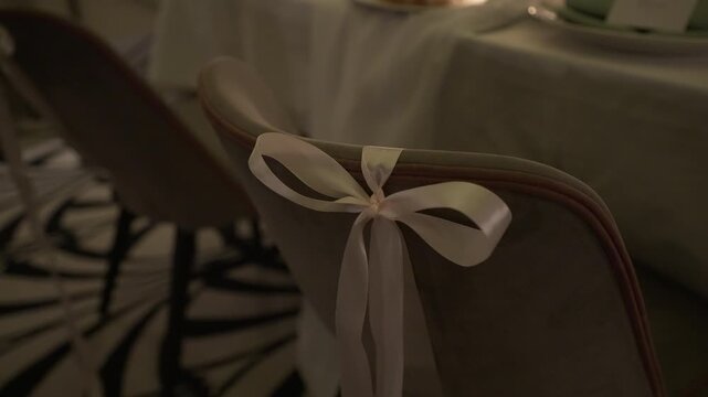 Banquet chair back with pale satin ribbon bow in dimly lit reception room, with chair, ribbon, bow, satin detail, showing upholstery, fabric, banquet, reception context