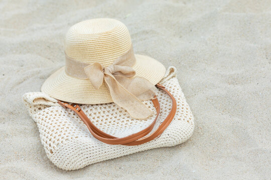 Straw sun hat featuring a ribbon sits atop a woven beach shopper bag on a light sandy surface, creating a relaxed summer vibe, perfect for beach outings or vacation