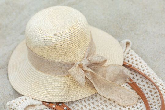 Straw sun hat with a light brown ribbon resting on a textured beach bag on sandy surface, ideal for summer outings or beach vacations