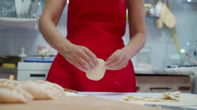 Close view of hands shaping filled dough on floured wooden board in home or restaurant kitchen, baker in red apron, with stainless steel backsplash, soft overhead light, and rustic culinary workflow