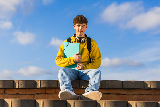 Young college student sitting on a wall holding a binder under blue sky