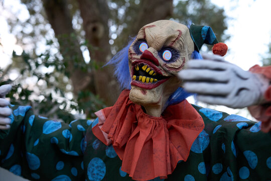 A menacing clown with spiral eyes reaches forward in a green polka-dot outfit. Blue hair, orange ruffle, and sharp yellow teeth complete the Halloween horror portrait.