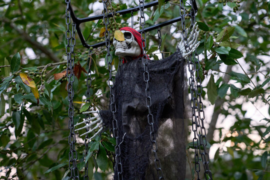 Close-up of a skeleton pirate in a hanging cage with plastic chains and a black shroud. The red bandana, weathered skull, and surrounding foliage form a creepy outdoor Halloween scene.