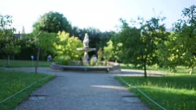 Blurred park scene with shallow bokeh and soft focus on a gravel path toward a fountain and statue; overlay backplate copyspace calm.