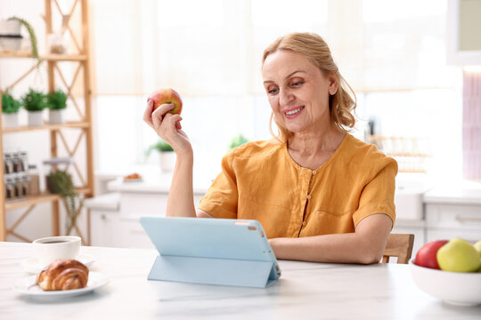 Smiling senior woman with apple having videocall by tablet at white marble table in kitchen