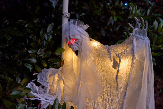 A white ghost decoration glows among leaves with bright red eyes at night. Draped fabric and warm lights create a spooky Halloween yard display.