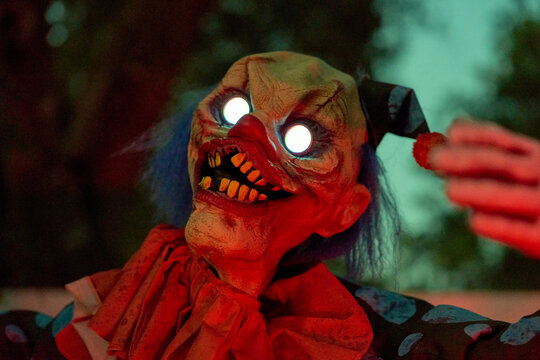 Closeup portrait of a creepy clown with bright glowing eyes and a red nose. Yellow teeth and harsh red light create a scary Halloween mood.