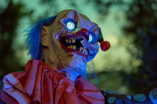Closeup of a scary clown with bright spiral eyes and blue hair. Red nose, yellow teeth, and eerie lighting make a spooky Halloween display.