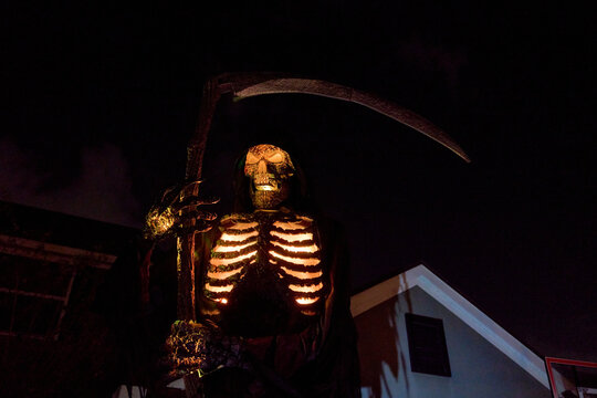 Hooded skeleton reaper glows with orange ribs and eyes at night. Dramatic Halloween yard display with a large curved scythe.