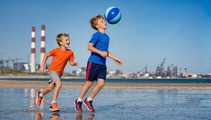 Two children play with a colorful ball in the foreground while blurred industrial smokestacks and coastal buildings rise against a bright blue sky. © ginettigino