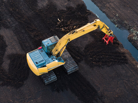 Overhead drone view of a yellow tracked excavator operating on a dark peat bog and clearing drainage channels in a wetland